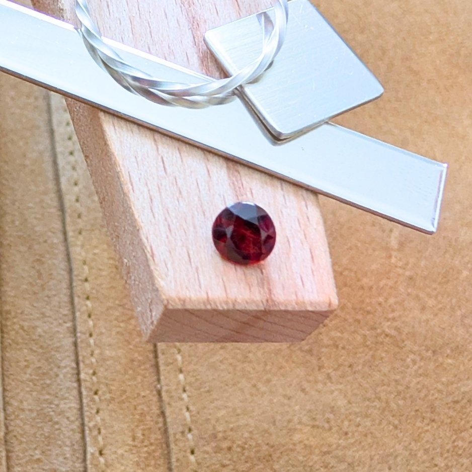 The loose faceted red garnet round is being displayed on a wooden jewellers bench peg with some sterling silver sheet and wire.