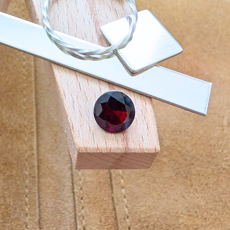 The loose faceted red garnet round is being displayed on a wooden jewellers bench peg with some sterling silver sheet and wire.