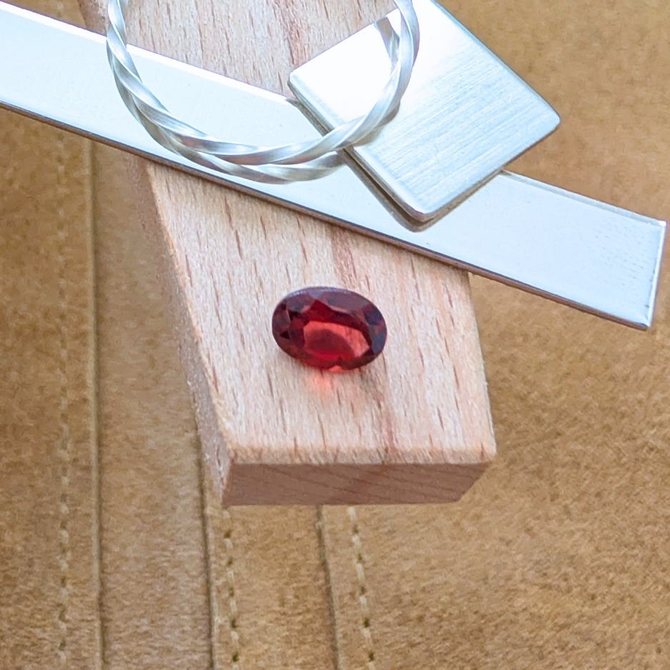 The loose faceted red garnet oval is being displayed on a wooden jewellers bench peg with some sterling silver sheet and wire.