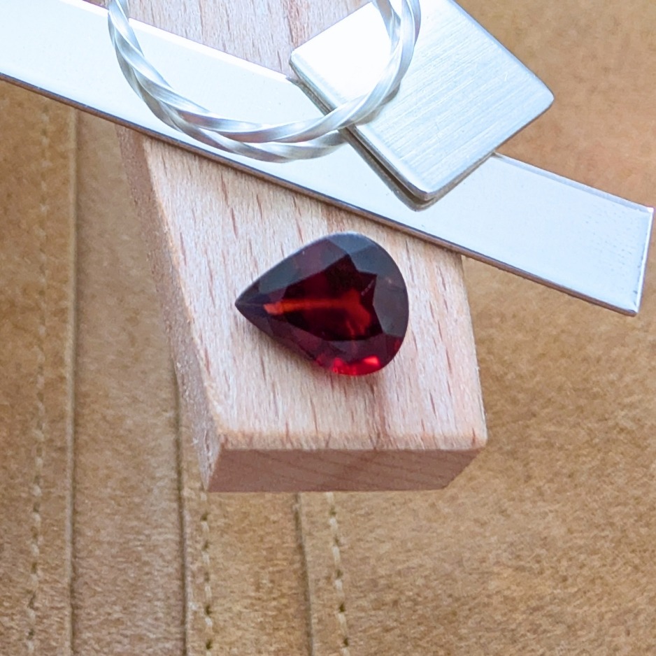 The loose red garnet teardrop is being displayed on a wooden jewellers bench peg with some sterling silver sheet and wire.