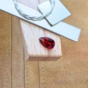 The loose red garnet teardrop is being displayed on a wooden jewellers bench peg with some sterling silver sheet and wire.