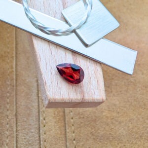 The loose red garnet teardrop is being displayed on a wooden jewellers bench peg with some sterling silver sheet and wire.