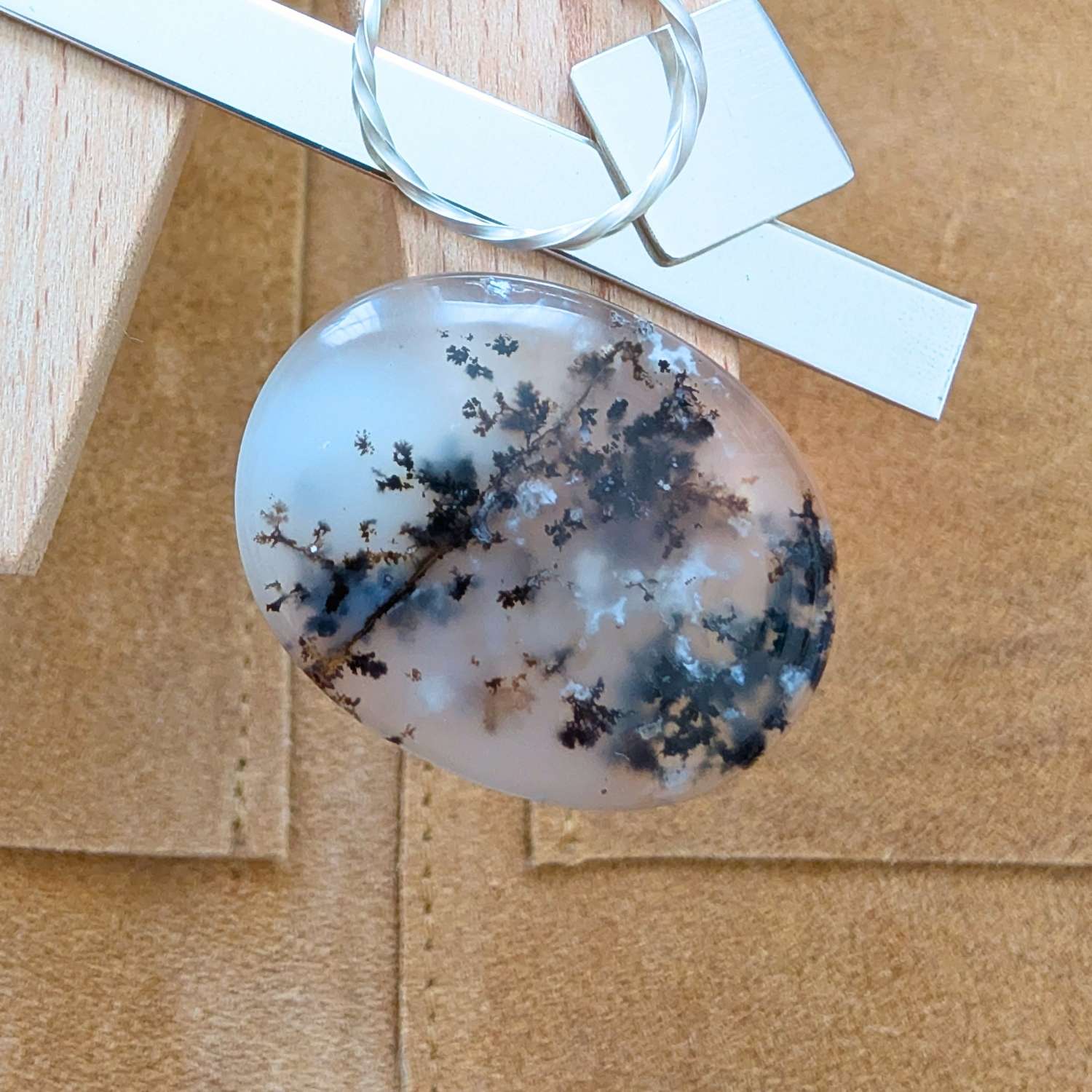 The loose dendritic opal cabochon is being displayed on a wooden jewellers bench peg with some sterling silver sheet and wire.