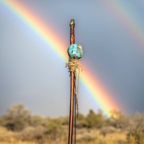 The top half of a wooden bow with a turquoise stone against a vibrant rainbow in a cloudy sky above a blurred, arid landscape, conveying tranquility.