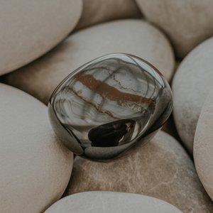 A smooth, reflective black Obsidian stone rests on a bed of matte white stones. The surface has light, organic patterns, creating a serene and balanced vibe.
