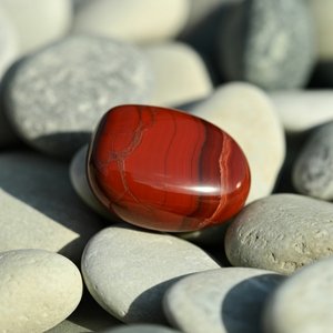 A smooth, polished red jasper stone with natural stripes sits on a bed of light gray pebbles, creating a striking contrast