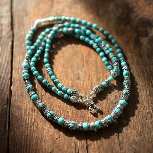 Turquoise and silver beaded necklace on rustic wood, with ornate patterns. Sunlight highlights intricate details, conveying a vintage, artisanal feel.