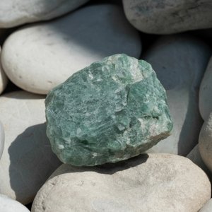 A rough green feldspar stone rests on smooth, white pebbles. The contrast highlights the stone's texture.